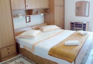 A neatly made bed with a white and beige bedspread, a gold throw blanket, and two pillows. There is a wooden wardrobe to the left and a small wooden cabinet above the bed. - House Marus (Omis)