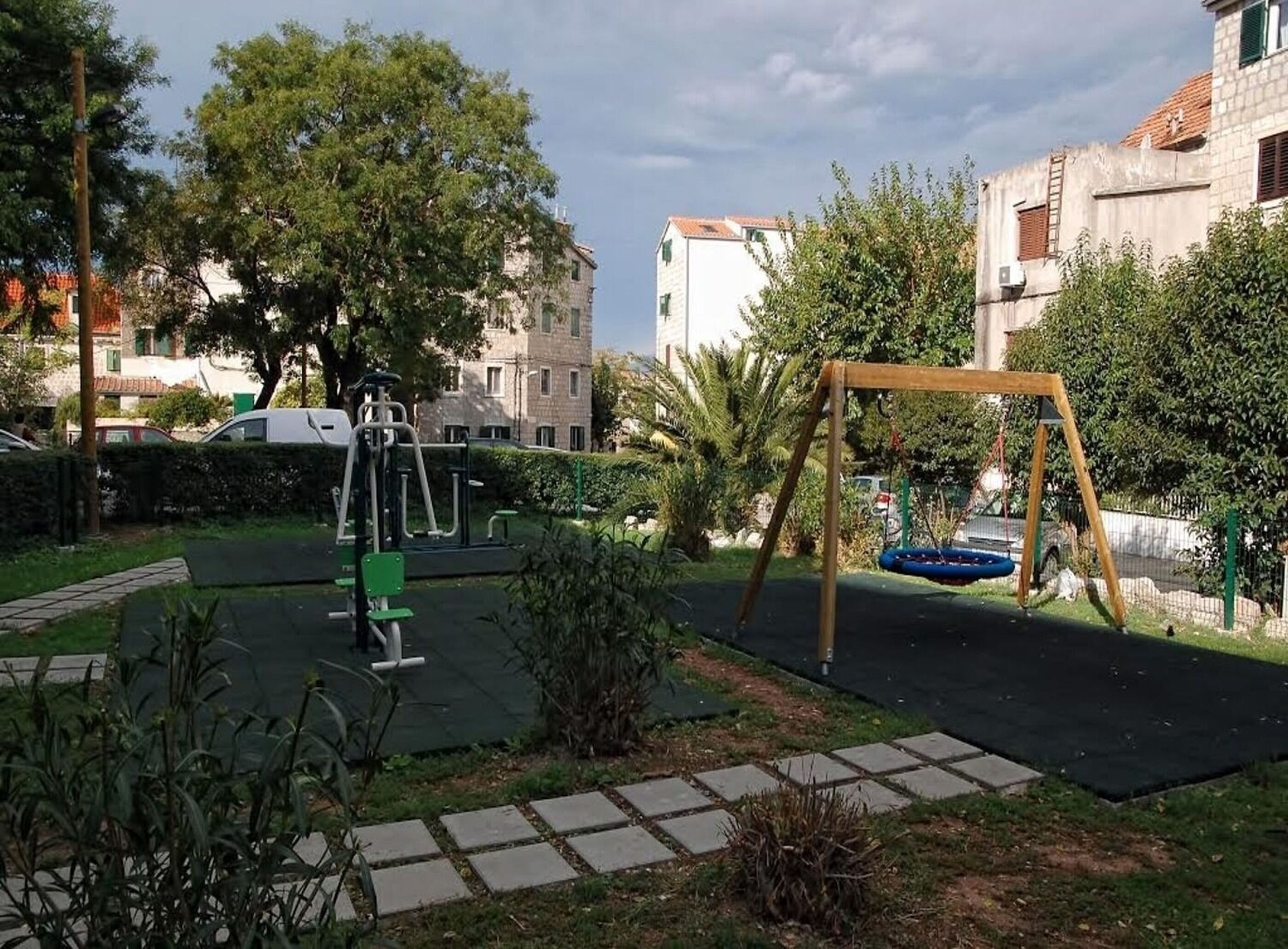 A playground with swings and exercise equipment, surrounded by greenery and buildings.