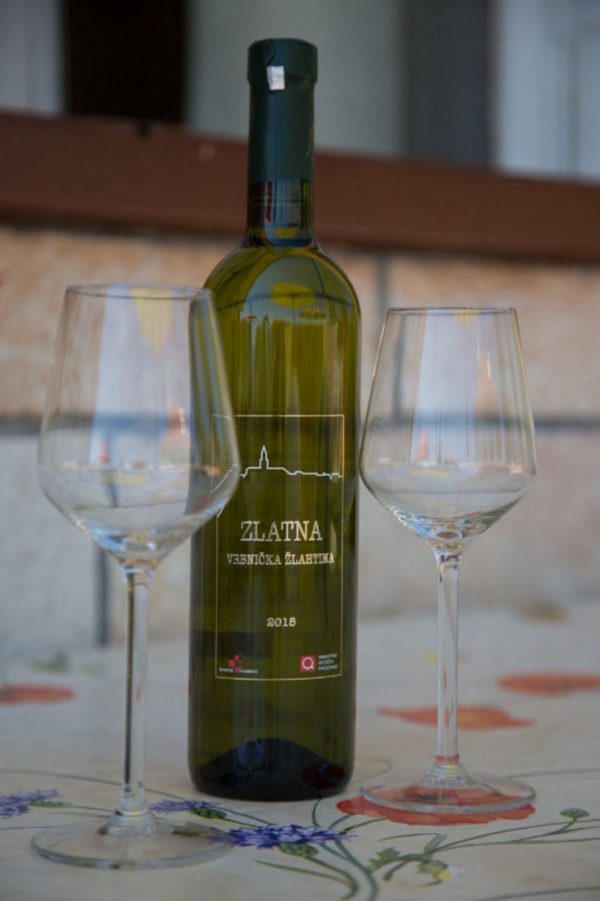 A bottle of ZLATNA wine with two empty wine glasses on a table with a floral pattern.