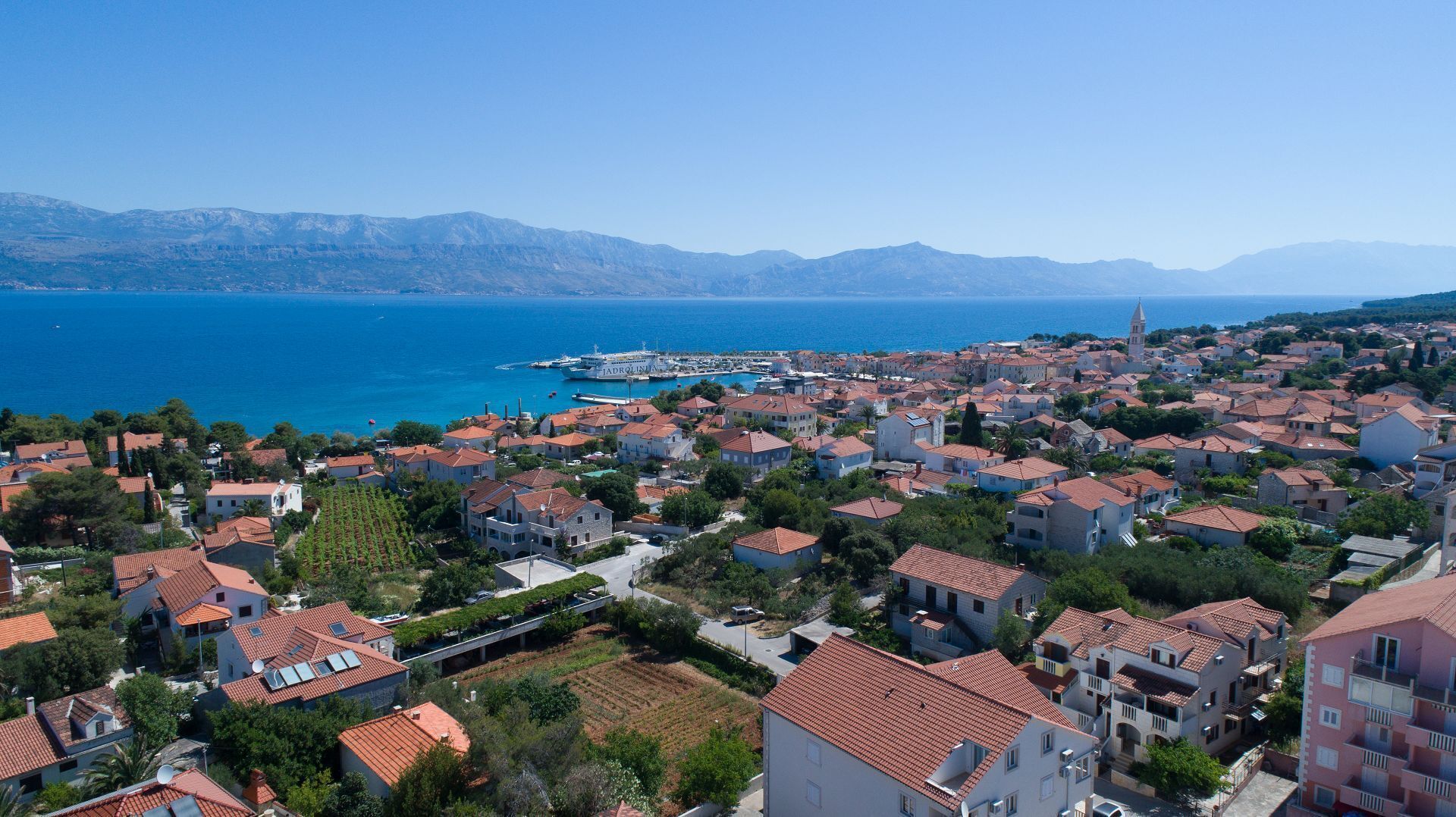 A coastal town with terracotta-roofed buildings, a marina, and a clear view of the sea and mountains in the distance.