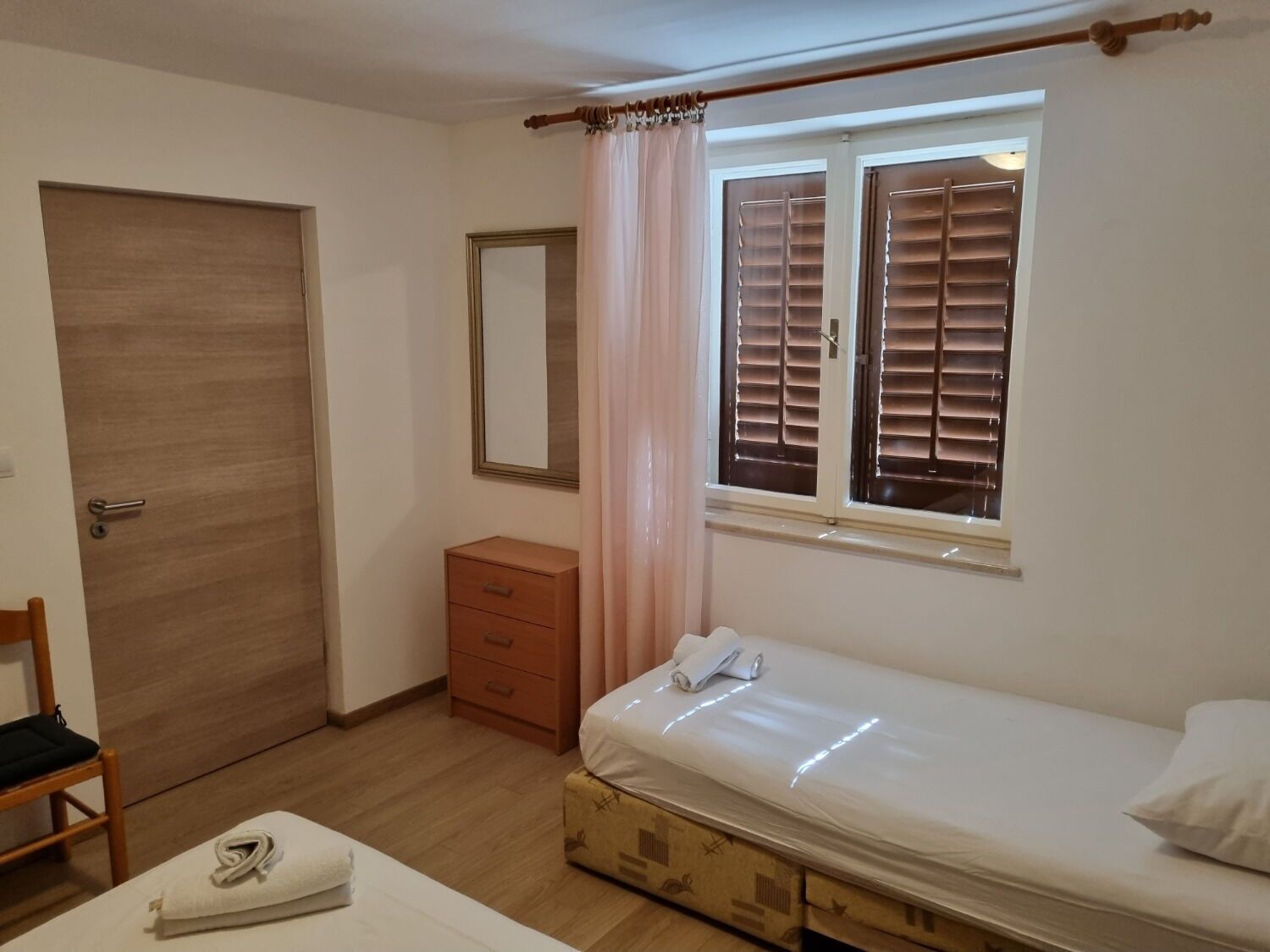A bedroom with a single bed, a wooden wardrobe, a small bedside table, a mirror, and a window with blinds.