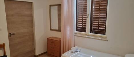A bedroom with a single bed, a wooden wardrobe, a small bedside table, a mirror, and a window with blinds.