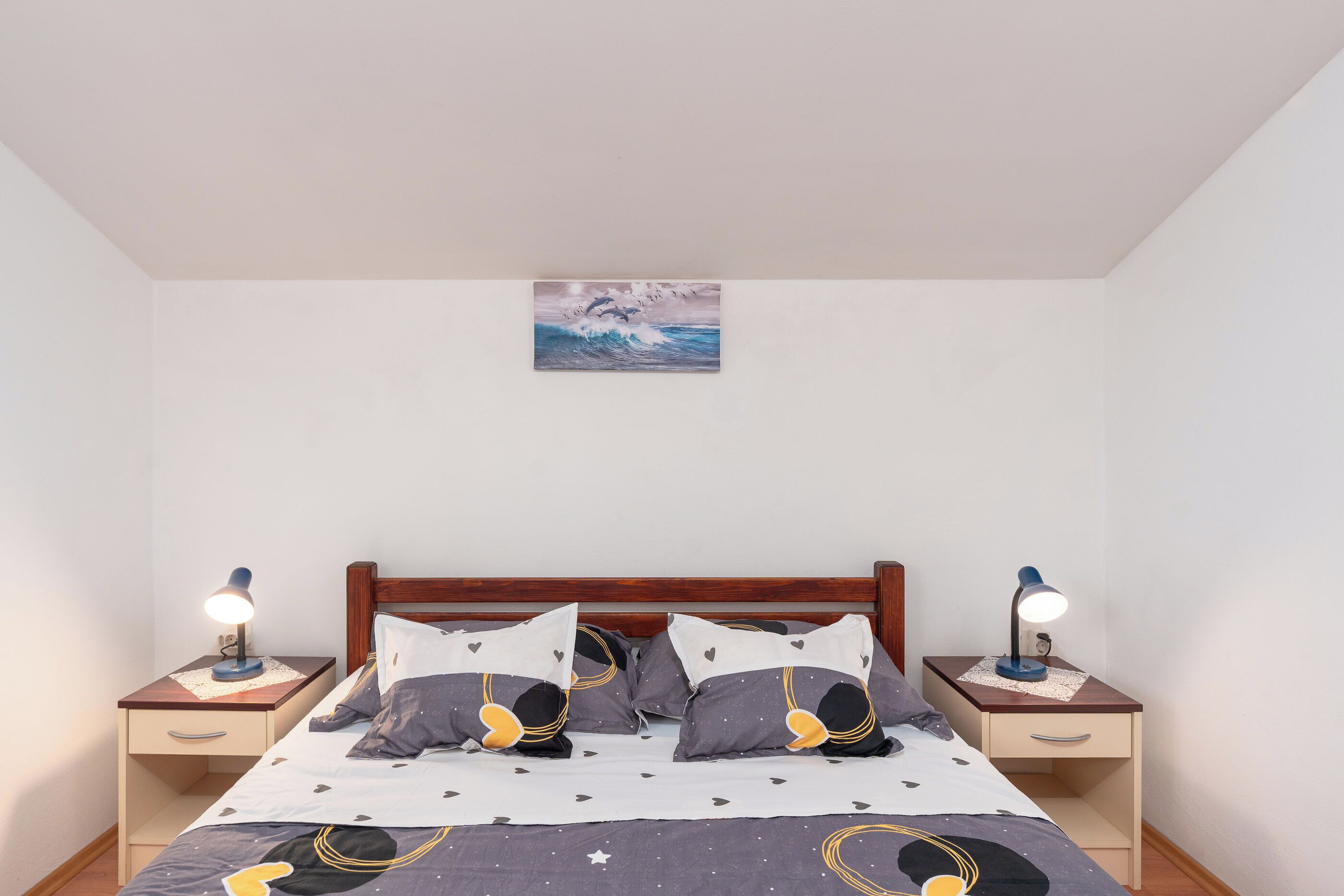 Quarto com cabeceira de madeira, cama com colcha estampada em formato de estrelas, duas mesas de cabeceira com luminárias e um quadro na parede.
