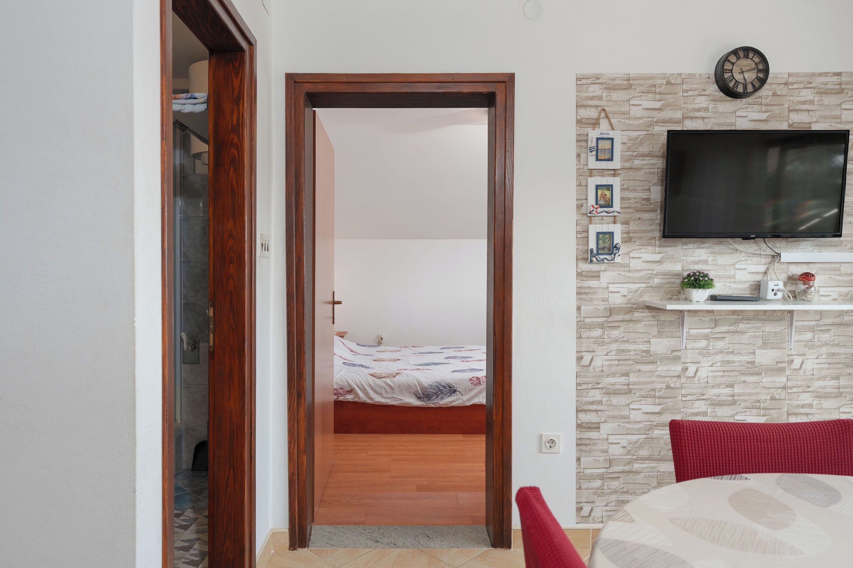 Quarto com porta de madeira que dá para um banheiro, cama com colcha florida, televisão fixada em uma parede com textura de pedra e uma cadeira vermelha.