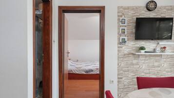 Quarto com porta de madeira que dá para um banheiro, cama com colcha florida, televisão fixada em uma parede com textura de pedra e uma cadeira vermelha.