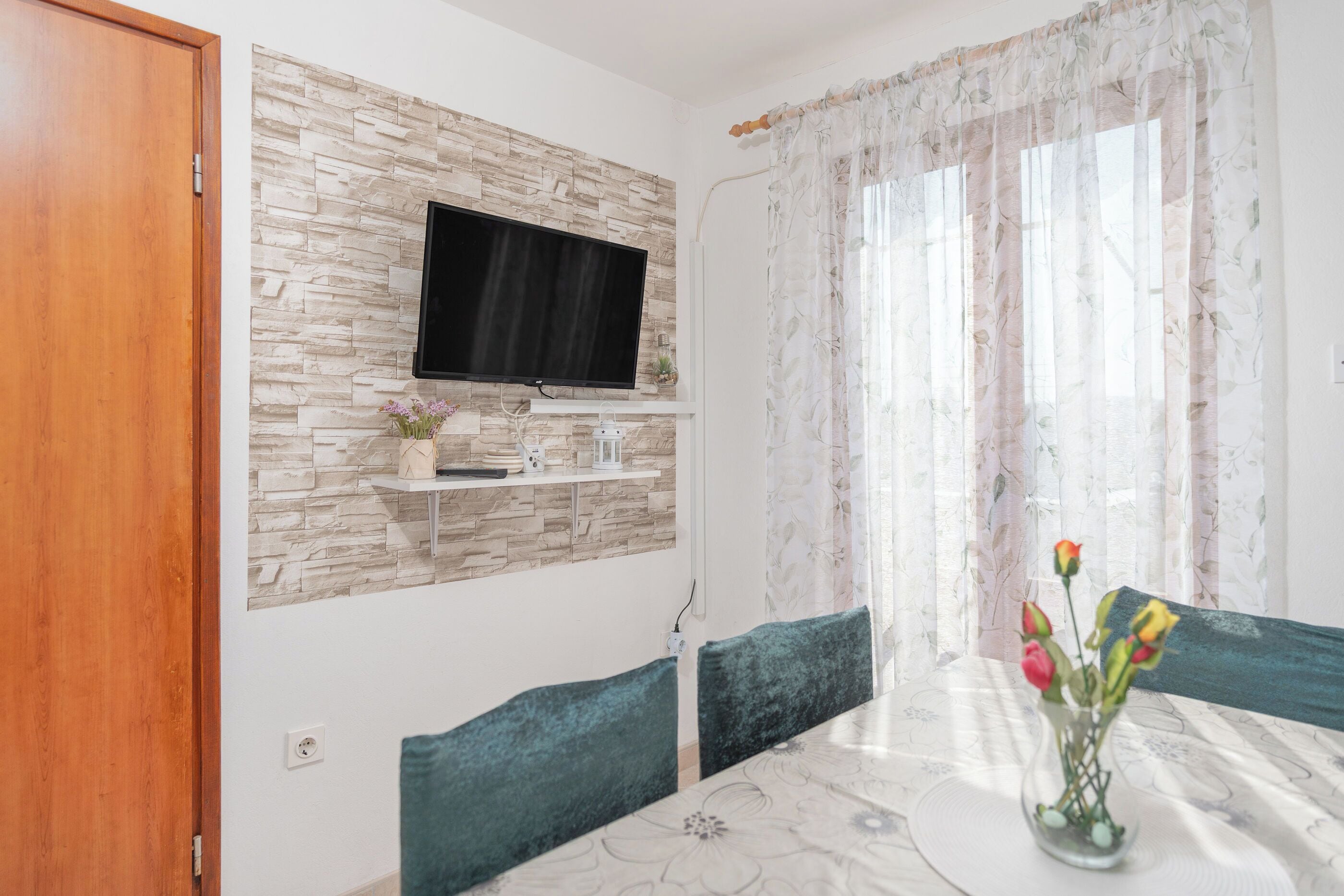 Uma sala de jantar com uma mesa, cadeiras, um vaso com flores, uma televisão fixada em uma parede de pedra e uma janela com cortinas leves.