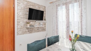 A dining area with a table, chairs, a vase with flowers, a television mounted on a stone wall, and a window with sheer curtains. - Apartments Darko (Vir)