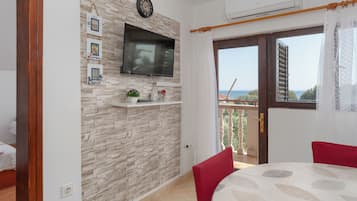 Quarto com parede de pedra, televisão montada, pequena prateleira com objetos decorativos, varanda com vista e área de jantar com mesa e cadeiras.
