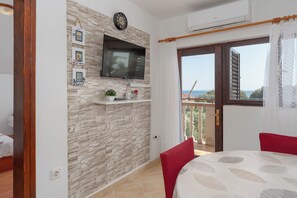 A room with a stone wall, a mounted TV, a small shelf with decorative items, a balcony with a view, and a dining area with a table and chairs. - Apartments Darko (Vir)