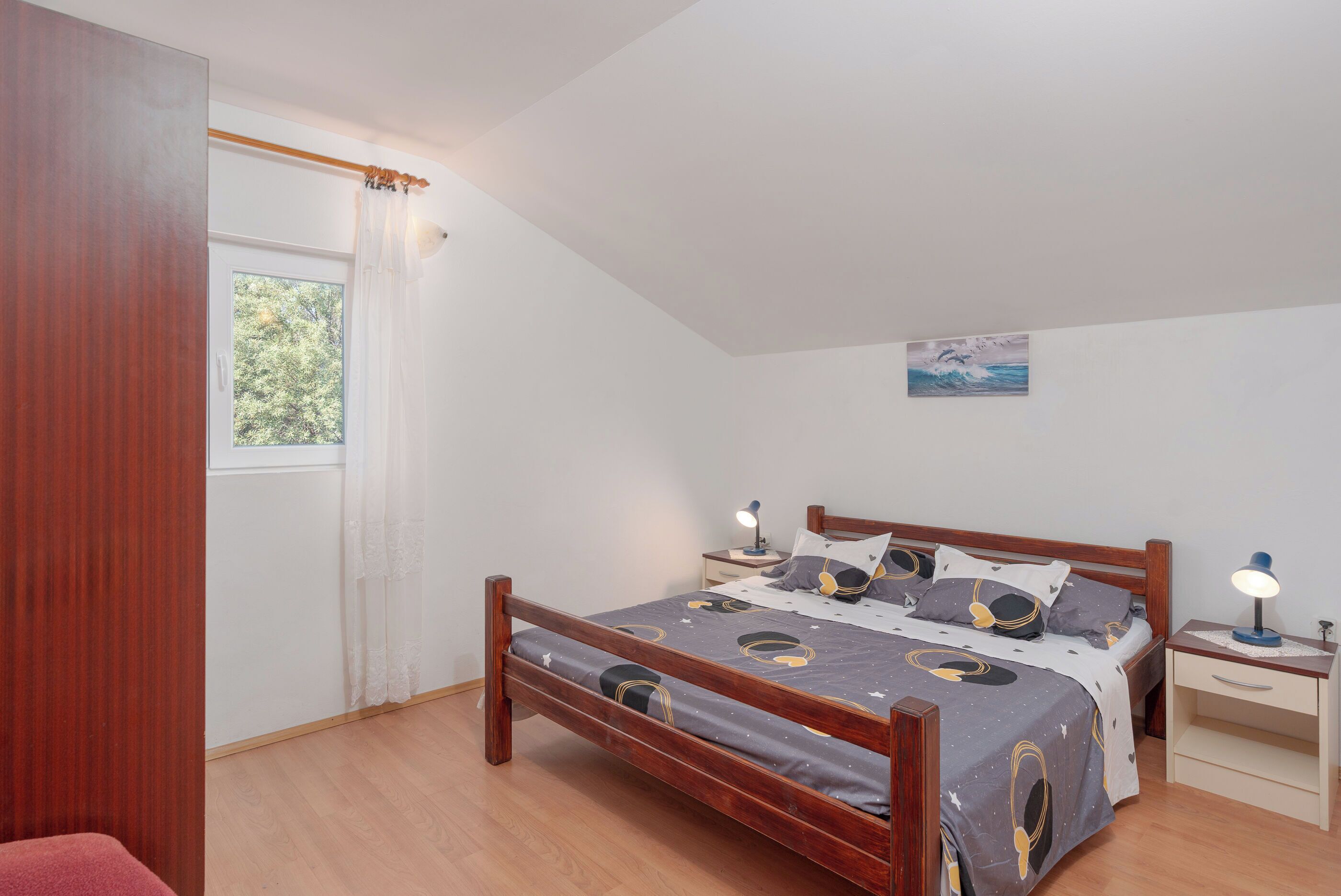 A bedroom with a wooden bed, a nightstand with a lamp, a window with curtains, and a painting on the wall.