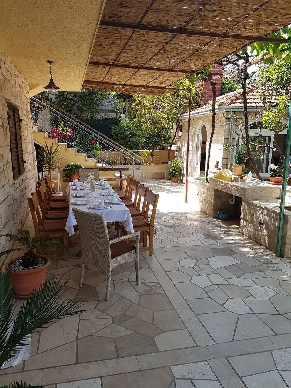 Um pátio externo com mesa posta para refeições, cercado por ladrilhos de pedra e plantas em vasos.