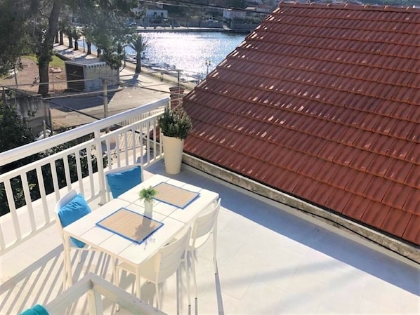 Un balcon avec une table et des chaises blanches, un pot de fleurs et une vue sur une rivière et des bâtiments.