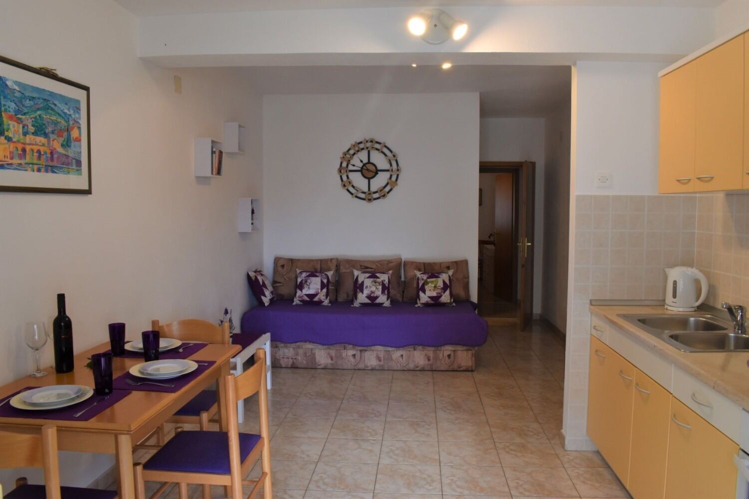 A compact living space with a dining area, a sofa with cushions, a kitchen with a sink and cabinets, and a wall-mounted clock.