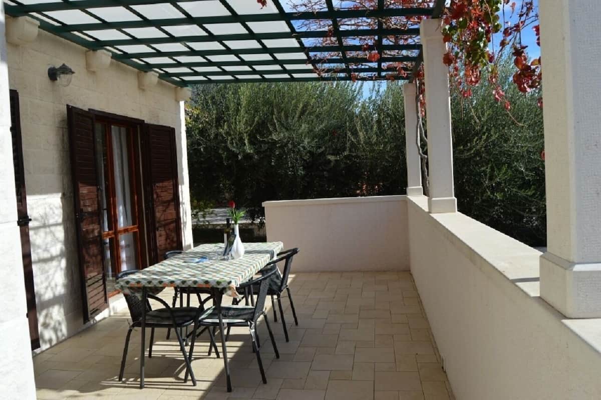 An outdoor patio with a table and chairs, a pergola, and a view of trees and a building.
