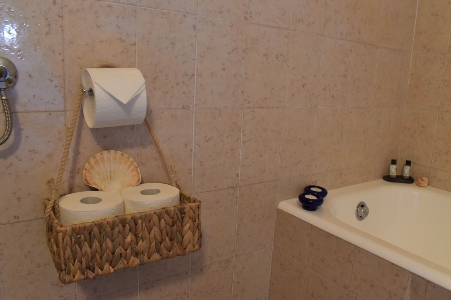 Une salle de bain avec un panier en osier contenant du papier toilette et un coquillage, une baignoire avec deux bouteilles et une pomme de douche.