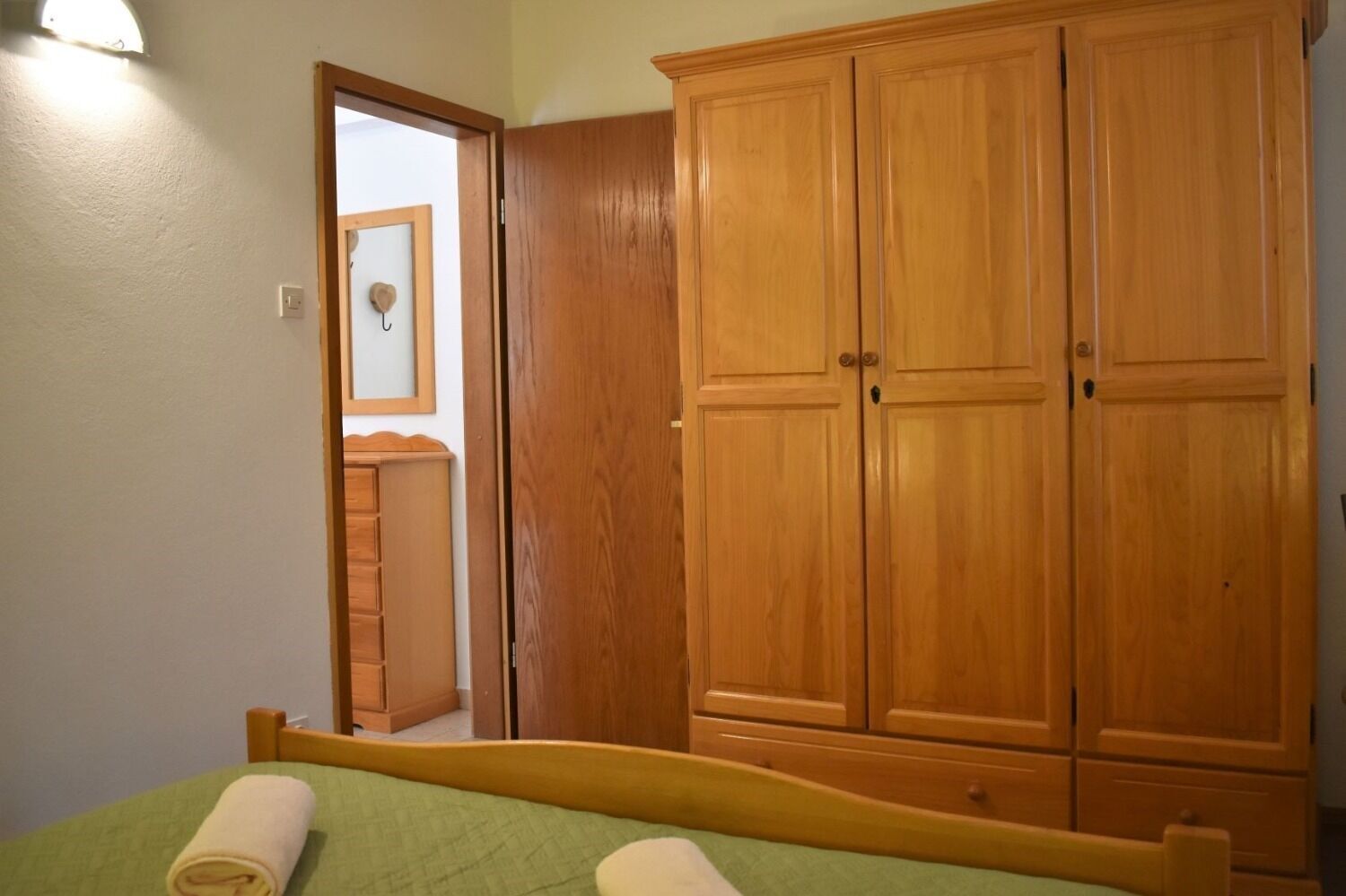 Een slaapkamer met een houten garderobe, een bed met een groene sprei en een spiegel aan de muur.