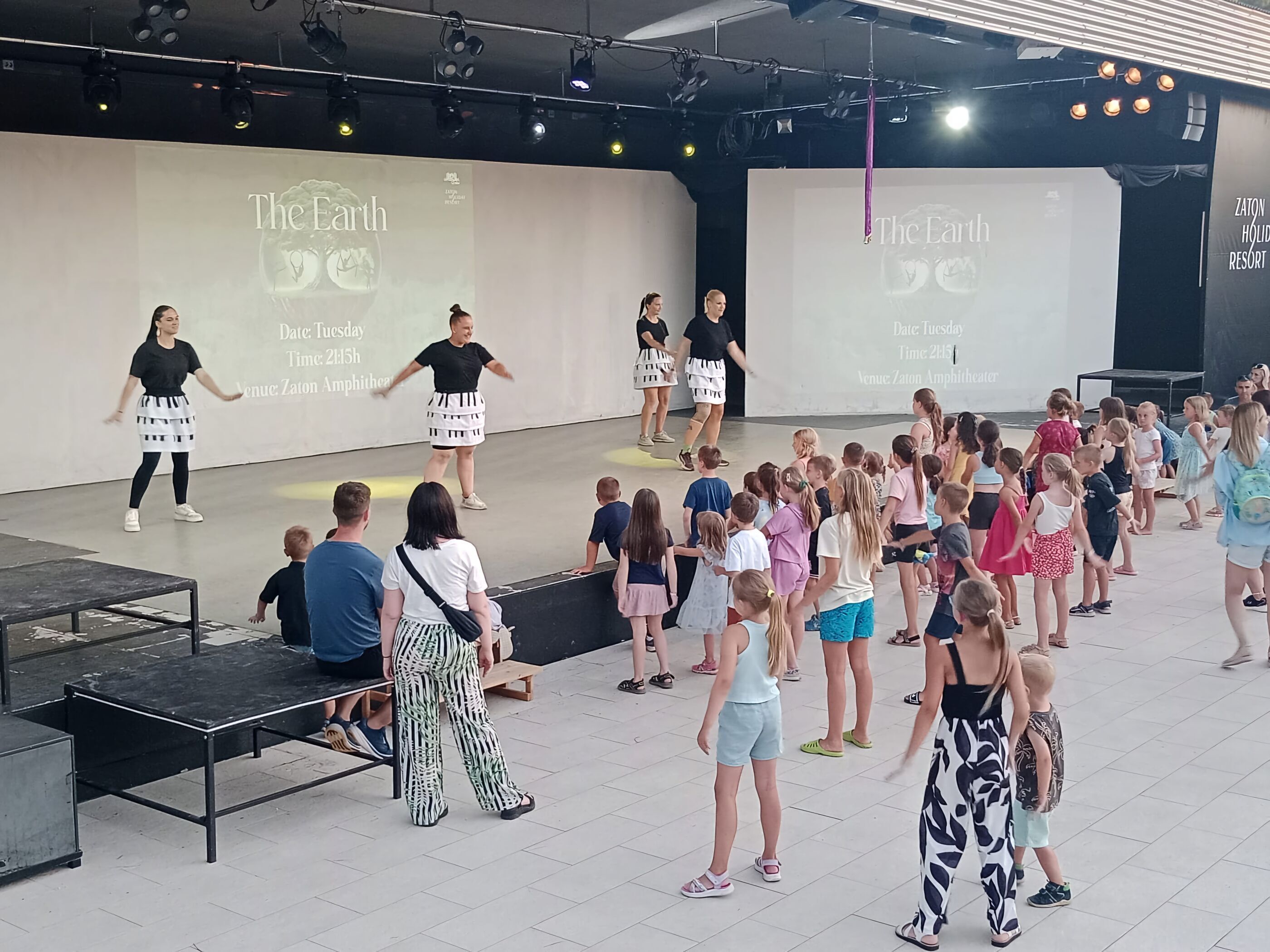 A stage with a group of people performing, a crowd of children watching, and a banner reading "The Earth" in a large indoor venue.