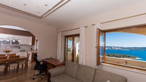 Een moderne woonkamer met een bank, een eethoek en een keuken. Een groot raam biedt uitzicht op zee.
