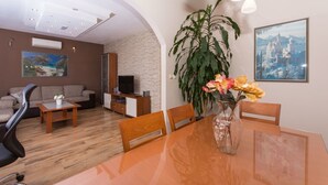 Een moderne woonkamer met een houten vloer, een bank, een televisie, een eettafel met stoelen en een grote plant.