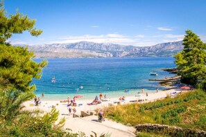 A beach with clear blue water, people swimming and sunbathing, and mountains in the background. - Apartment Pavlo (Postira)