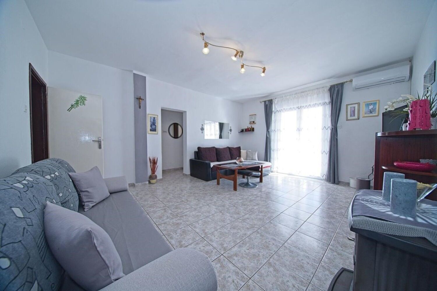 Ein geräumiges Wohnzimmer mit einer Couch, einem Couchtisch und einem Essbereich. Eine Tür führt in einen weiteren Raum und ein Fenster mit Vorhängen ist vorhanden.
