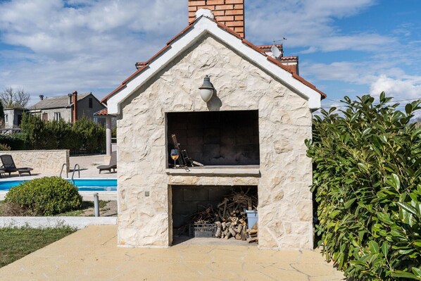 A stone-built outdoor barbecue area with a chimney, a glass door, and a pool in the background. - House Oasis Village Villa (Privlaka)