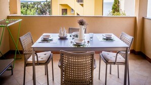 Uma área de refeições com uma mesa posta para quatro pessoas, cercada por cadeiras de vime, em uma varanda com vista para um edifício amarelo e o mar.