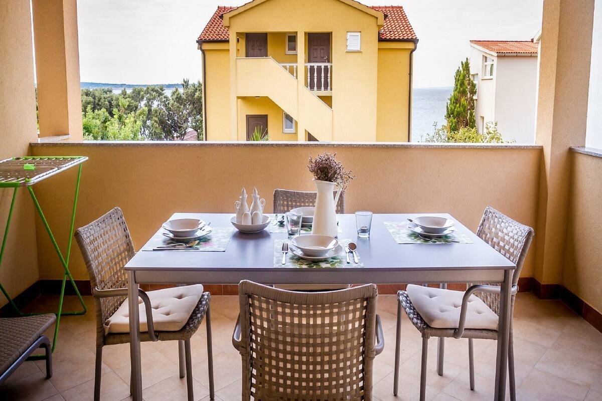 A dining area with a table set for four, surrounded by wicker chairs, on a balcony with a view of a yellow building and the sea.