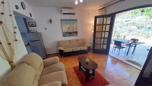 Sala de estar com sofá, mesa de centro e um quadro na parede. Há uma porta que dá para uma área externa com mesa e cadeiras.