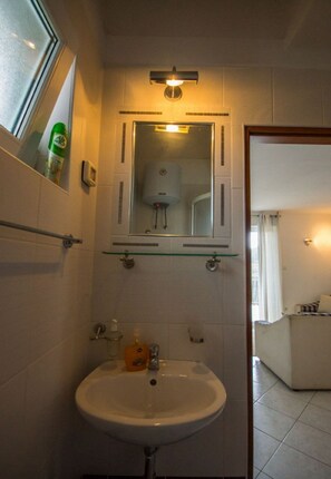 Et badeværelse med en glashylde over et hvidt håndvaskarrangement, et spejl med lys over, og et toilet i baggrunden.