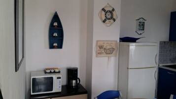 Une cuisine compacte équipée d’un four à micro-ondes, d’une machine à café et d’un réfrigérateur. On y trouve une étagère bleue ornée d’objets décoratifs et un tableau encadré accroché au mur.