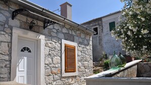 A stone building with a white door and shutters, a small window, and a garden with potted plants and a tree. - Apartments Oasis (Sali)