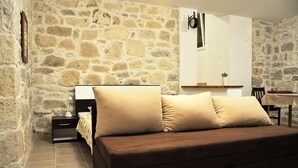 A cozy room with a stone wall, a brown sofa with beige cushions, a bedside table, and a small desk with a chair. - Apartments Oasis (Sali)