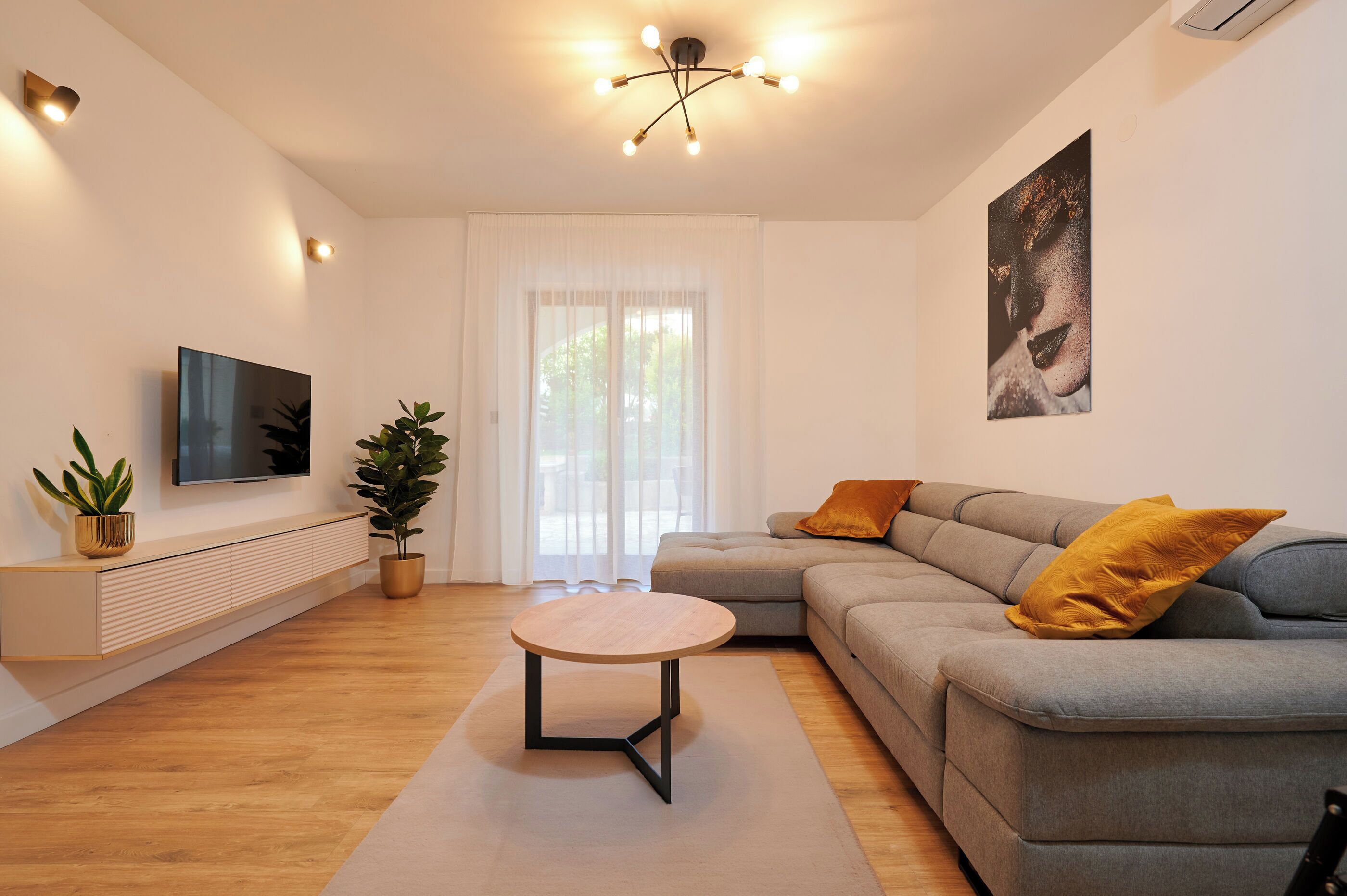 Un salon moderne avec un canapé gris, une table basse ronde, un téléviseur à écran plat fixé au mur et une grande fenêtre ornée de rideaux légers.