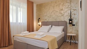 A neatly made bed with white linens and a beige throw, a bedside table with a lamp, and a wall with a textured pattern. - Apartment Natali (Okrug)