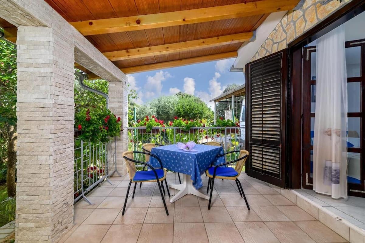 A covered patio with a table and chairs, surrounded by a stone wall and a garden with red flowers.