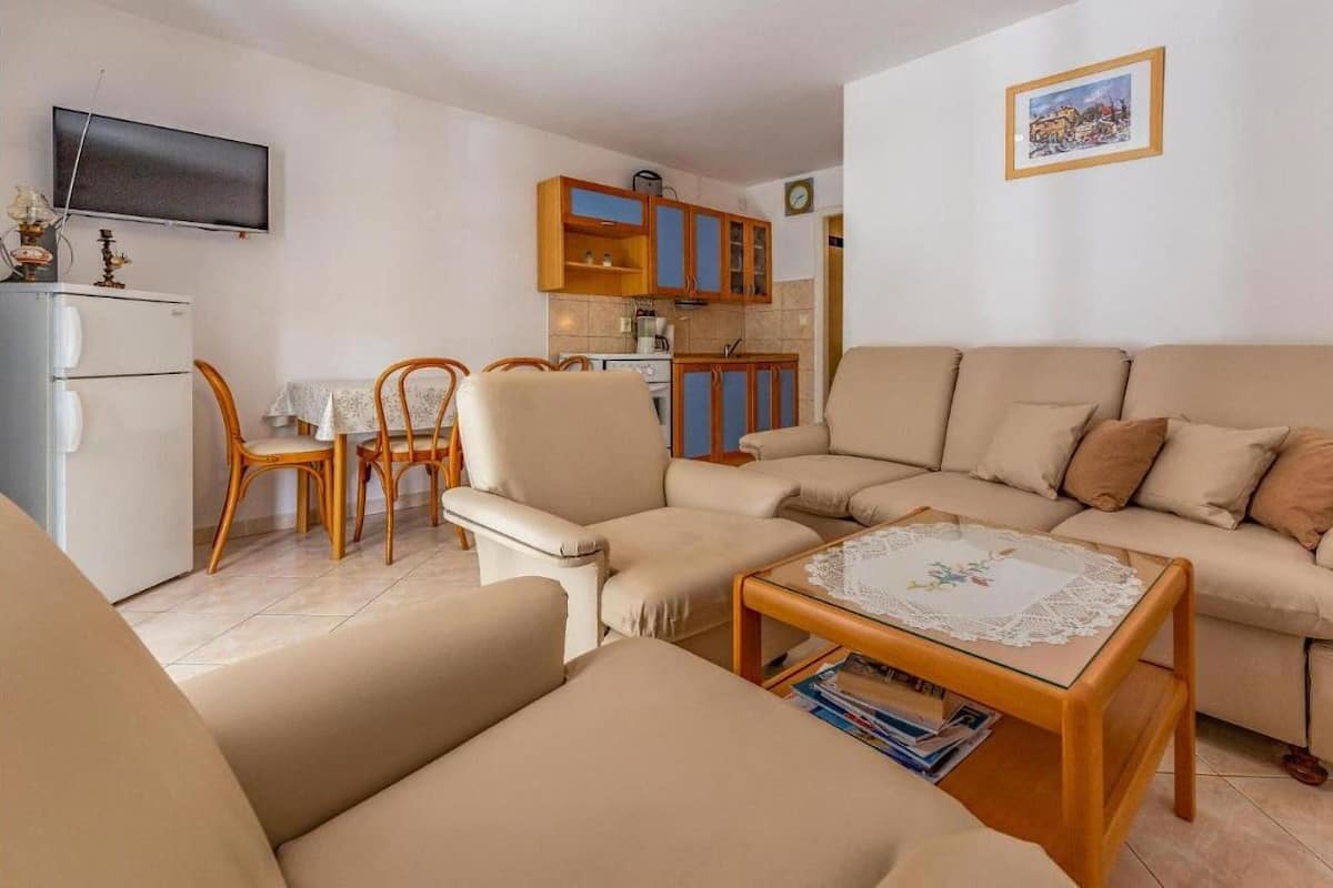 A living room with a sofa, chairs, a coffee table, a TV, a kitchen area with cabinets, and a clock on the wall.