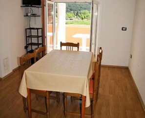 Ein Essbereich mit Tisch und Stühlen, einem Fernseher auf einem Regal und Blick auf eine grüne Landschaft durch eine offene Tür.
