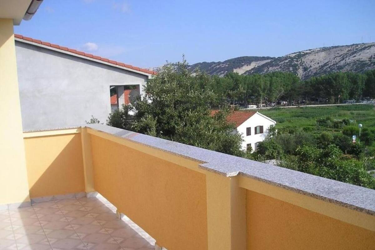 A balcony with a yellow wall and a tiled floor, overlooking a green landscape with trees and a white building.