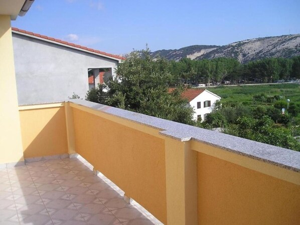 Ein Balkon mit gelber Wand und Fliesenboden, mit Blick auf eine grüne Landschaft mit Bäumen und einem weißen Gebäude.