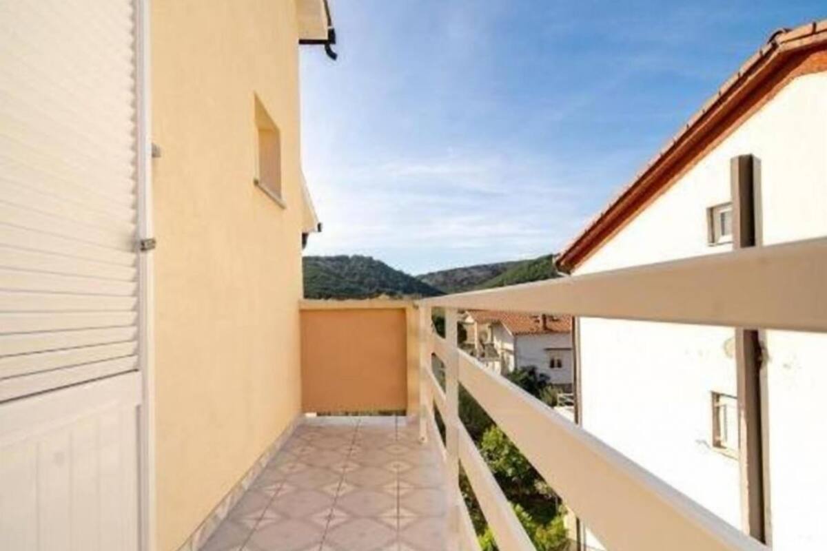 A narrow balcony with a tiled floor, a white railing, and a view of neighboring buildings and hills.