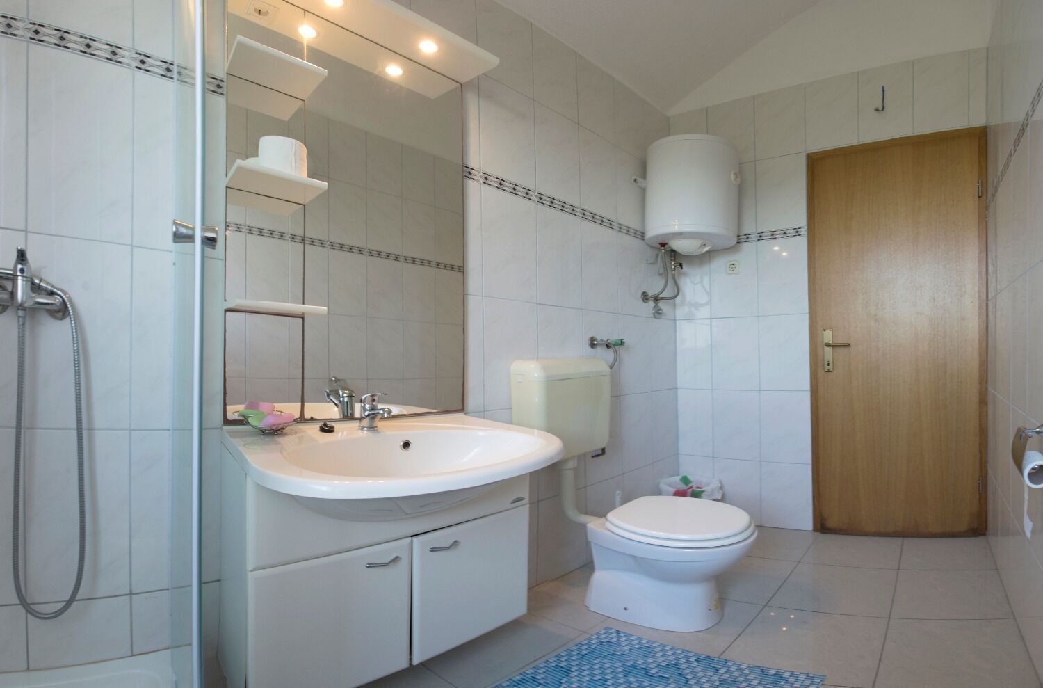 A bathroom with a white sink, toilet, and shower. There is a mirror above the sink and a wooden door to the right.