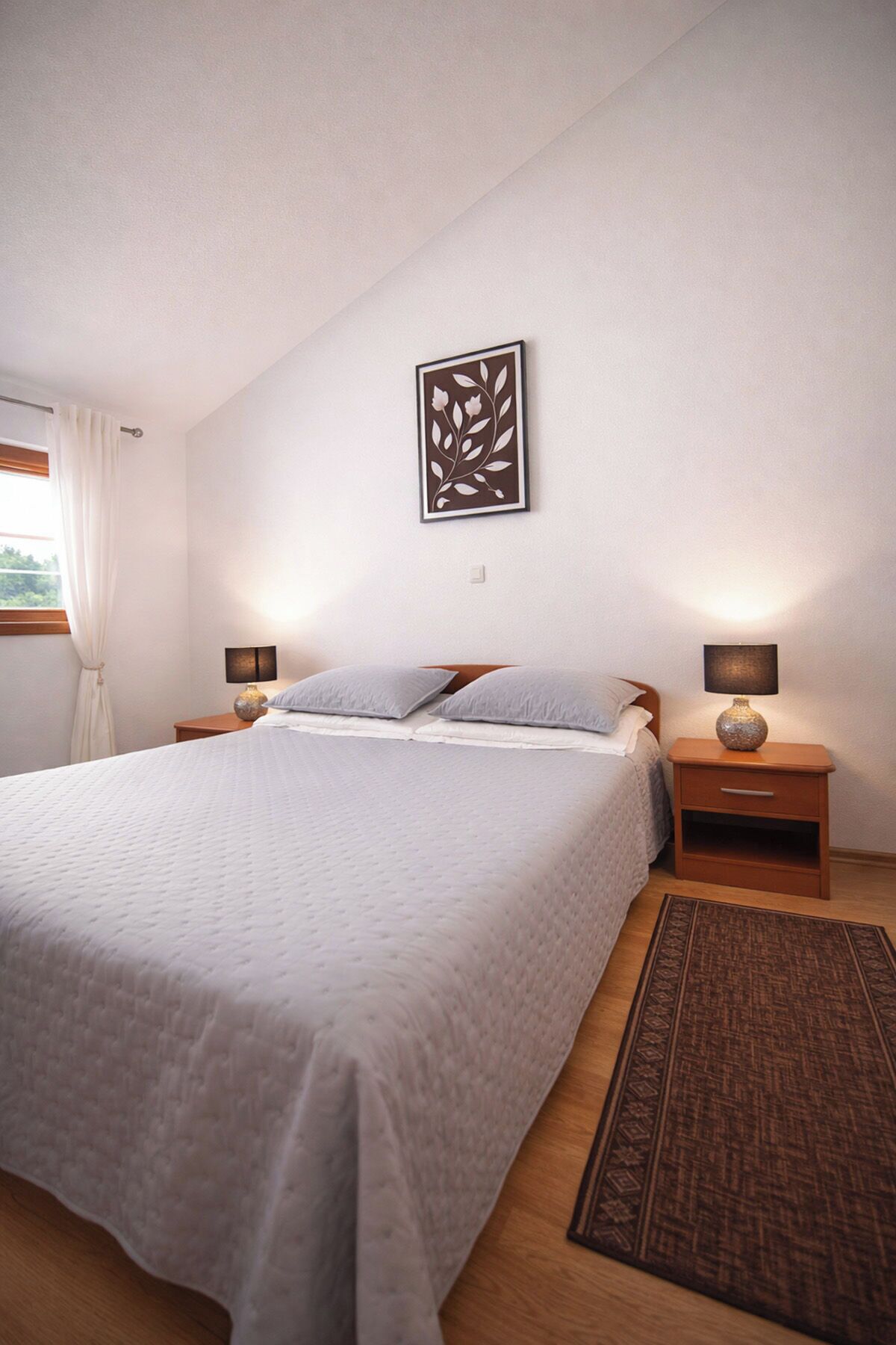 Quarto com cama, duas mesas de cabeceira, uma obra de arte na parede, janela com cortinas e um tapete.