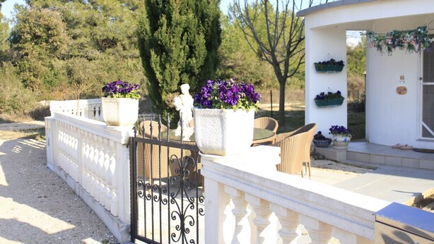 A gated entrance with a white balustrade, potted purple flowers, wicker furniture, and a white building with a door and windows. - Apartments Marin (Biograd na Moru)
