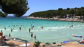 Ein Strand mit klarem türkisblauem Wasser, Menschen beim Schwimmen und Sonnenbaden, sowie eine felsige Küstenlinie mit Bäumen.