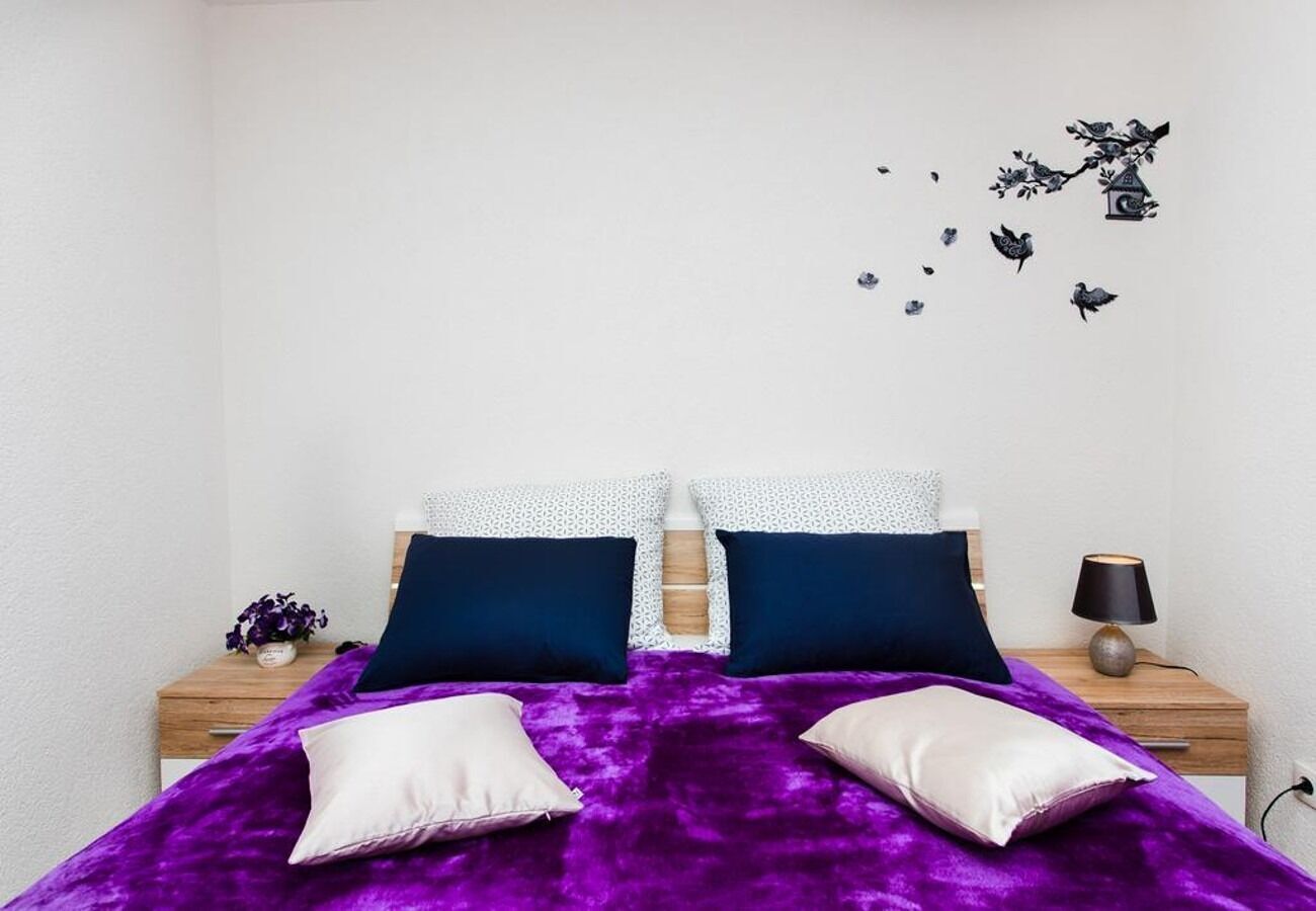 Una cama con manta y almohadas moradas, una mesita de noche con una lámpara, y un adorno en la pared con pájaros y una casa.