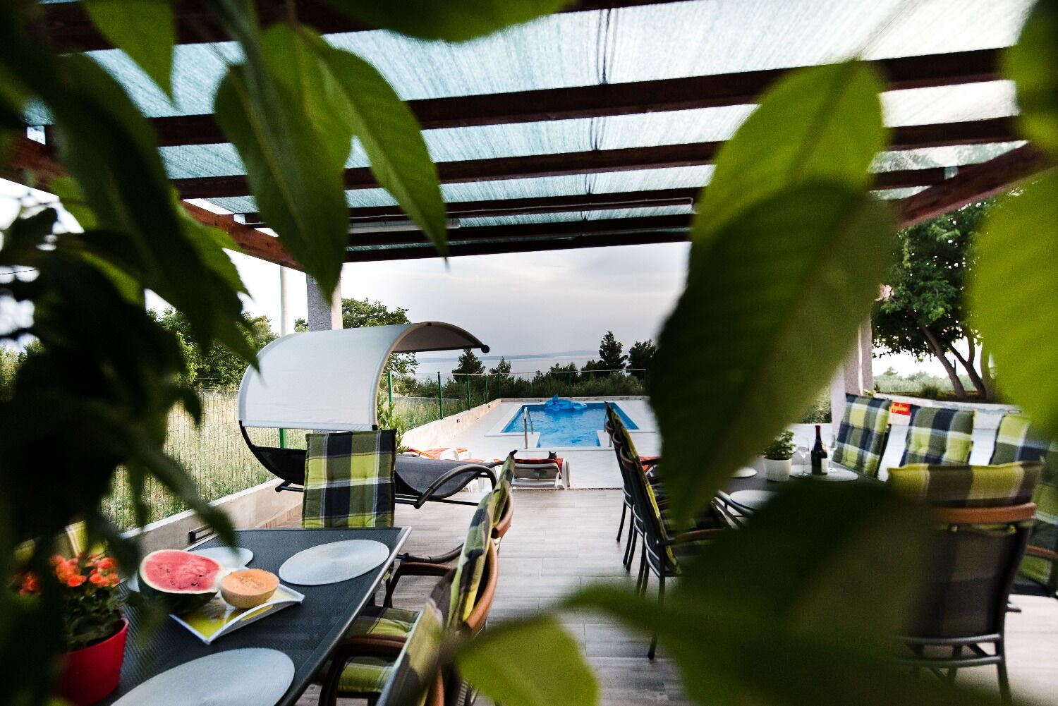 An outdoor dining area with a pool in the background, covered by a pergola.