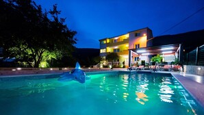 A swimming pool with a dolphin float, a covered outdoor seating area, and a building with multiple balconies in the background. - Apartment Mariska (Podstrana)