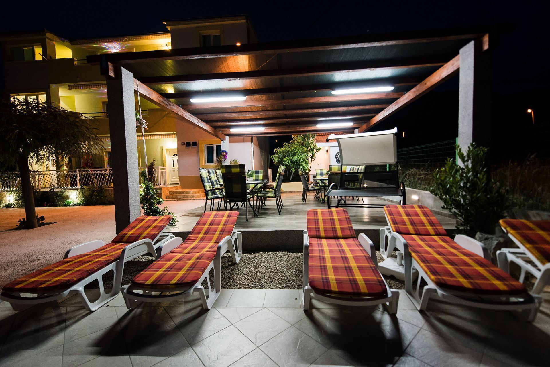 An outdoor patio with multiple seating areas, including sun loungers and chairs, under a covered structure with overhead lighting.