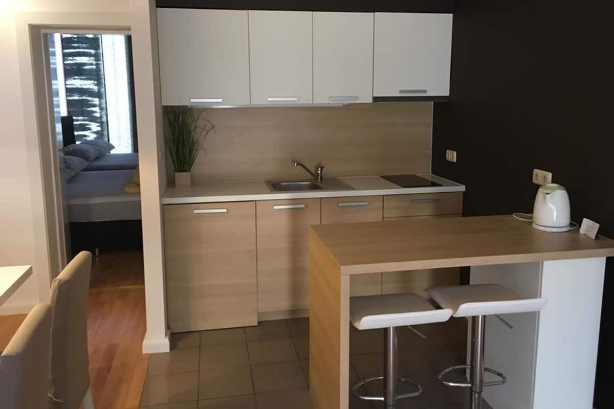 A modern kitchen with a built-in sink, a countertop, and a kettle. There is a window with a view of trees and a bed in the adjacent room.